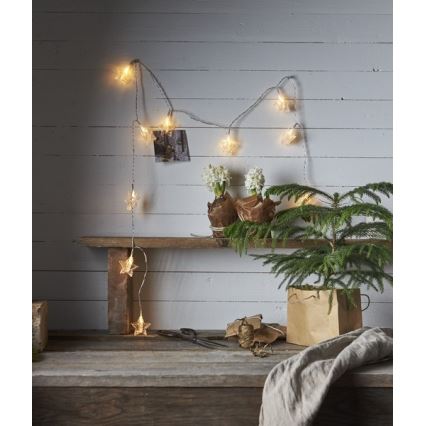 Svetielkový veniec s hviezdami svieti na drevenej stene. Veniec je umiestnený na poličke s kvetináčmi a vianočným stromčekom. Na stole pod poličkou sú nožnice a špagát.