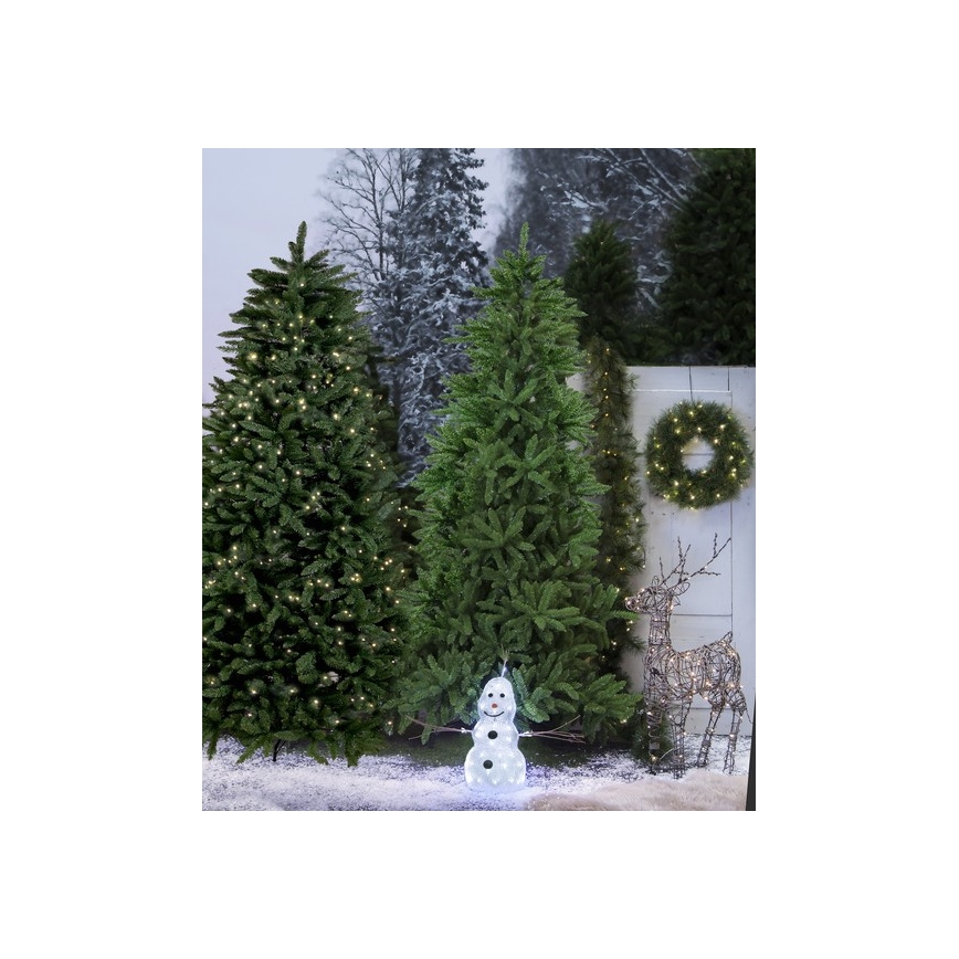 Vianočné stromčeky s osvetlením stoja pred bielou stenou. Popri nich je snehuliak s LED osvetlením, veniec a kovový jeleň. Snehová pokrývka je na zemi.