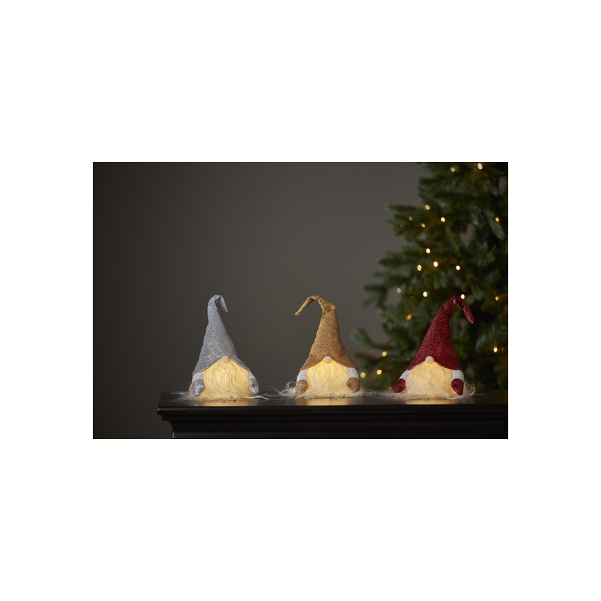 Tri trpaslíci s LED osvetlením sedia na krbovej rímse pred vianočným stromčekom. Trpaslíci majú čiapočky striebornej, zlatej a červenej farby a svetlé brady.