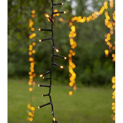 Elektrický svetelný reťaz s malými žltými LED diódami visí vonku na rozmazanom pozadí zelenej trávy a rozostrených žltých svetiel.