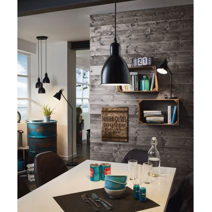 Čierna závesná lampa svieti nad jedálenským stolom. Na stene je drevený obklad. Na stole sú taniere, poháre a konzervy. Text na obrázku znie: HUILE D'OLIVES GARANTIE PURE Produit de Provence.