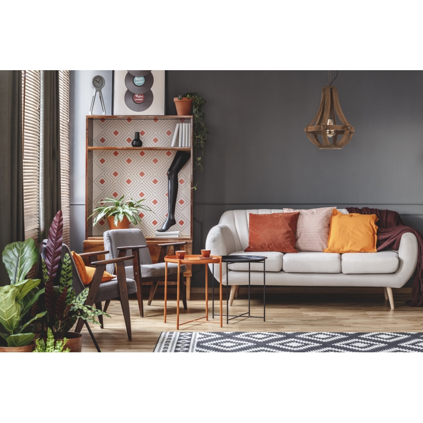 Drevená lampa visí v obývacej izbe. Svetlá sedačka s oranžovými a ružovými vankúšmi je umiestnená pred poličkou s geometrickým vzorom. V miestnosti sú dva kreslá a dva malé stolíky.