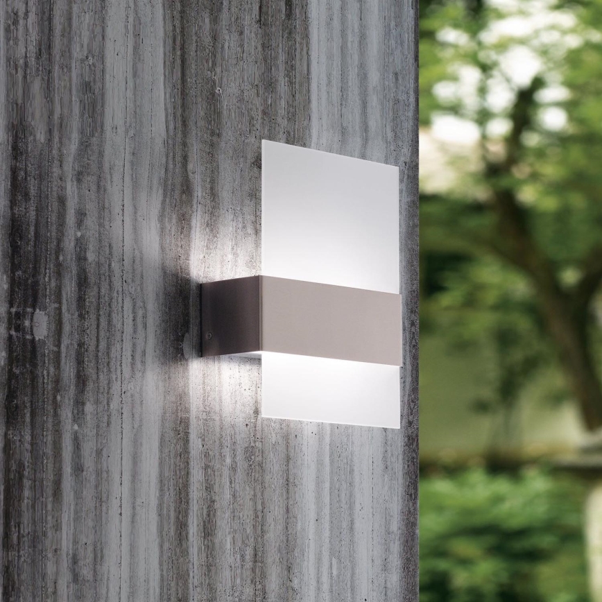 Nástenné svietidlo je pripevnené k vertikálnej betónovej stene. Svietidlo svieti. Na pozadí je rozmazané zelené záhradné prostredie.