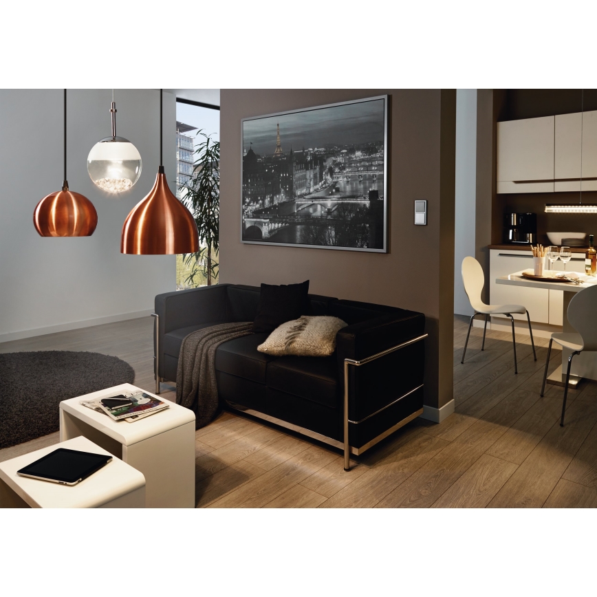 Tri visiace lampy, dve medené a jedna sklenená, svietia v obývacej izbe. Čierna kožená sedačka stojí pred obrazom mesta. V pozadí je vidieť kuchyňu s jedálenským stolom a stoličkami.