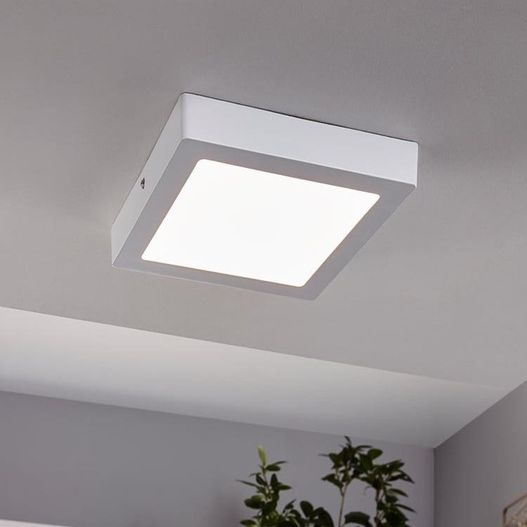 Štvorcové LED svietidlo je namontované na strope. Svietidlo je biele, s jemne presvitajúcim svetlom. V pozadí je vidieť časť miestnosti s rastlinou.