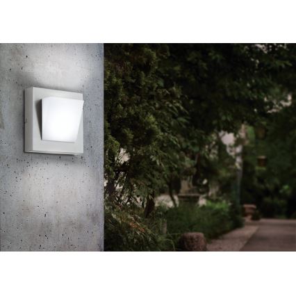 Nástené svietidlo svieti na betónovej stene v exteriéri. Svietidlo je štvorcové, sivé s bielym štvorcovým difúzorom. Pozadie tvorí zeleň záhrady a chodník.