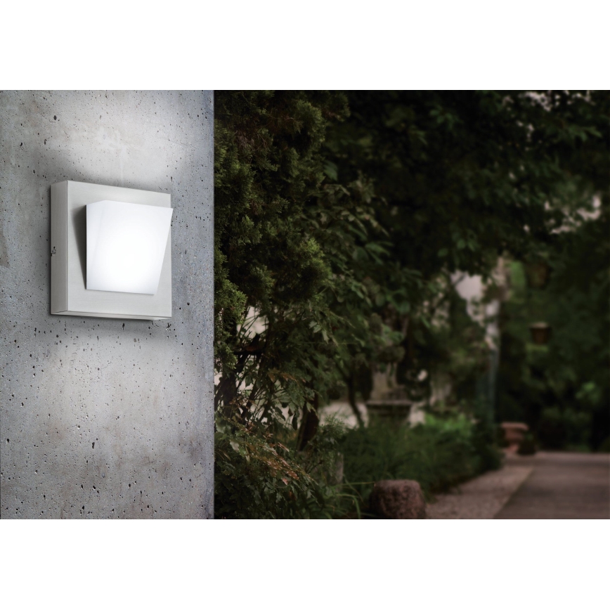 Nástené svietidlo svieti na betónovej stene v exteriéri. Svietidlo je štvorcové, sivé s bielym štvorcovým difúzorom. Pozadie tvorí zeleň záhrady a chodník.