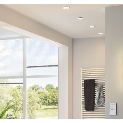 Svietidlá osvetľujú kúpeľňu. Na stene je radiátor s uterákmi. Veľké okno s žalúziami ponúka výhľad do parku. Na stene je vypínač.