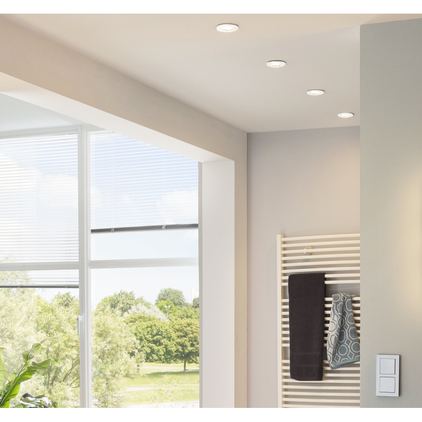 Svietidlá osvetľujú kúpeľňu. Na stene je radiátor s uterákmi. Veľké okno s žalúziami ponúka výhľad do parku. Na stene je vypínač.