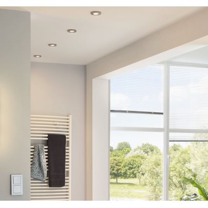 Štyri zapustené stropné svetlá svietia v kúpeľni. Na stene je radiátor s uterákmi. Veľké okno s žalúziami ponúka výhľad do parku.