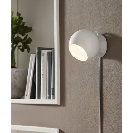 Biela nástenná lampa svieti. Je pripevnená na stene vedľa police s knihami a rastlinou v kvetináči.