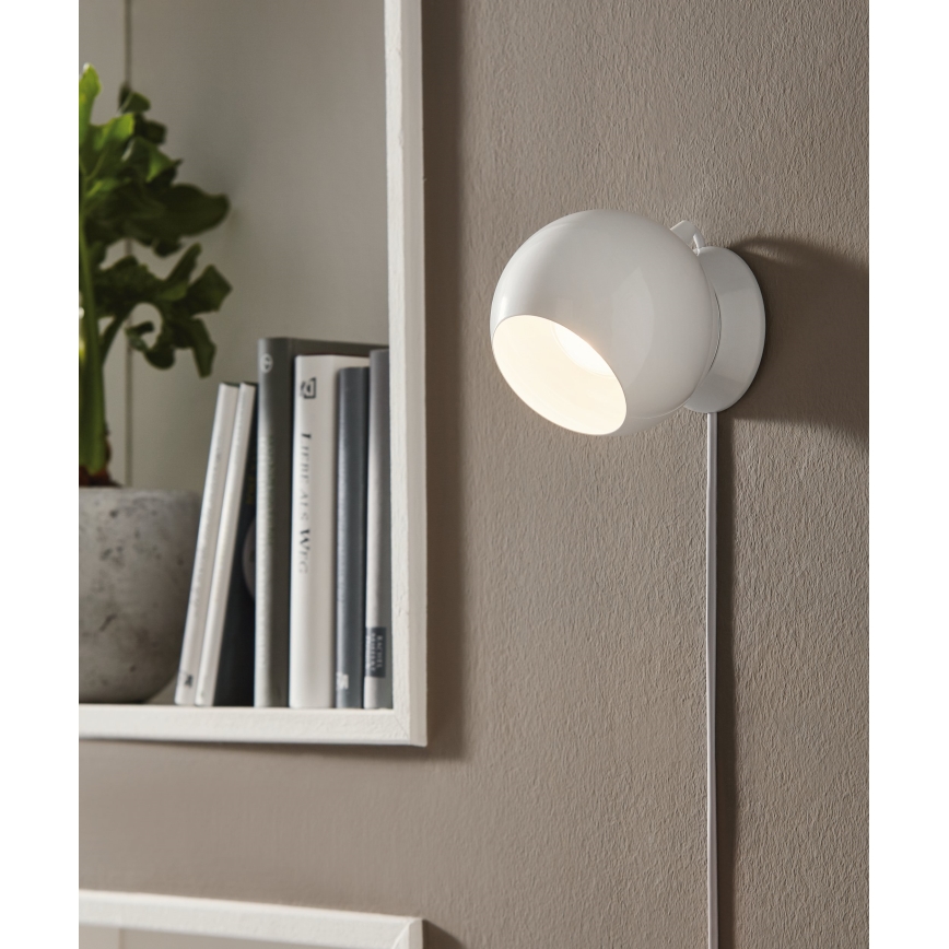 Biela nástenná lampa svieti. Je pripevnená na stene vedľa police s knihami a rastlinou v kvetináči.