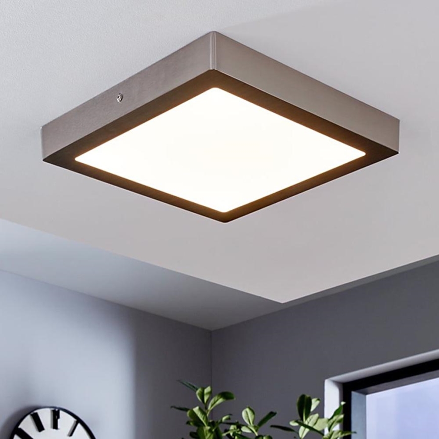 Štvorcové stropné svietidlo svieti v interiéri. Svietidlo má kovový rám a svetlý stred. Nachádza sa na svetlom strope v miestnosti so sivými stenami.