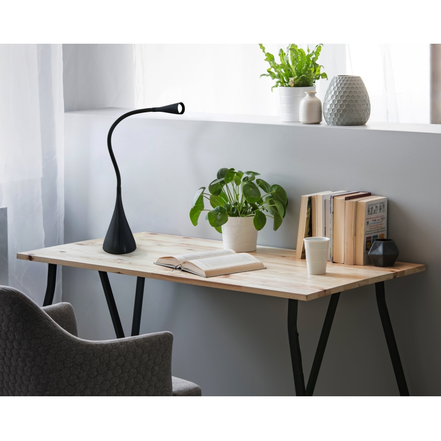Čierna stolová lampa s ohybným ramenom svieti na drevenom stole. Na stole leží kniha, kvetináč s rastlinou a hrnček. Na stene nad stolom sú ďalšie rastliny a vázy.