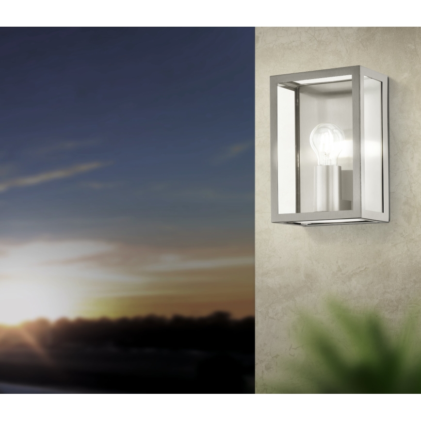 Nástená lampa svieti. Je umiestnená na betónovej stene s rastlinou v popredí. Na pozadí je západ slnka.