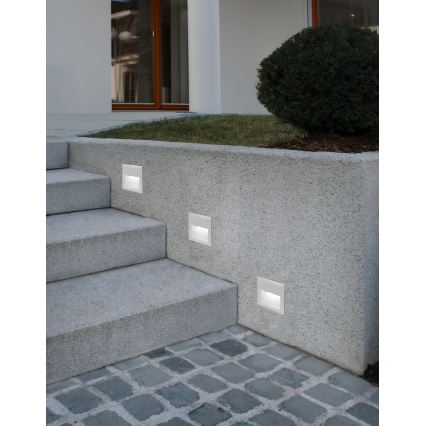 Tri nástenné svietidlá sú osadené do kamennej steny pri schodisku. Svietidlá sú zapnuté. Schodisko vedie k domu s oknami a kríkom.