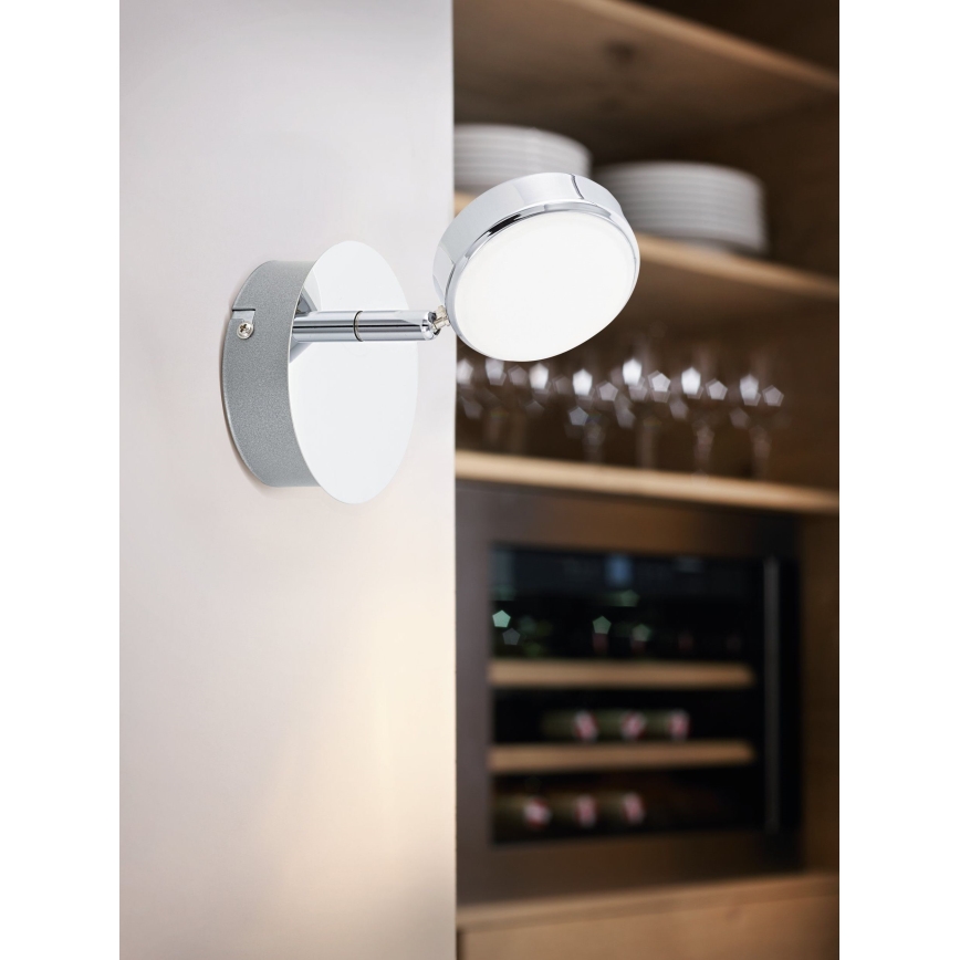 Nástenné svietidlo s otočnou hlavou je pripevnené na stenu. Svietidlo smeruje doprava. V pozadí sa nachádza kuchynská linka s vínnym chladiacim zariadením.