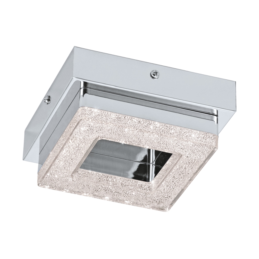 Stropné svietidlo s hranatým tvarom a chrómovým rámom svieti. Vnútorná časť je zdobená trblietkami. Svietidlo je zavesené v bielom pozadí.