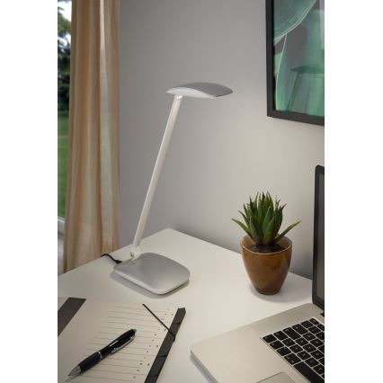 Stôlna lampa svieti na pracovnom stole. Nachádza sa na ňom aj notebook, zápisník, pero a kvetináč s rastlinou. Lampa je striebornej farby.
