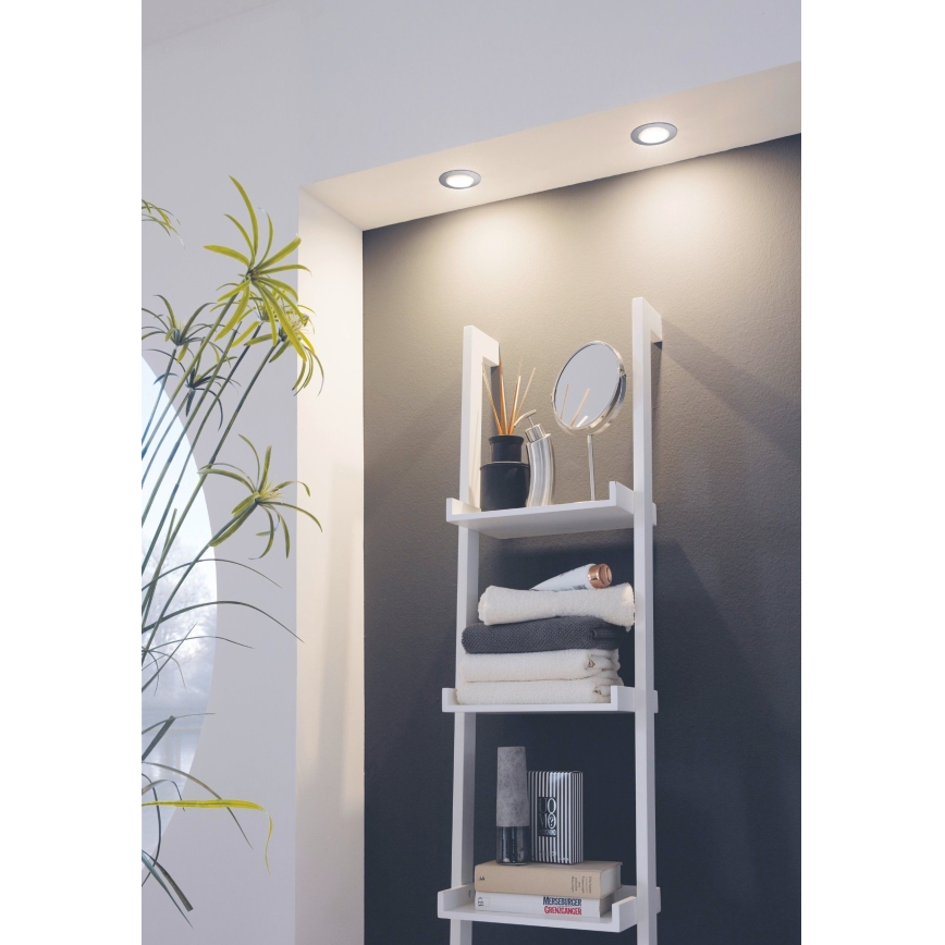 Biela polica s tromi policami – umiestnené sú na nej uteráky, knihy, dekorácie a zrkadlo. Polica je pripevnená na stene osvetlenej tromi stropnými svietidlami.