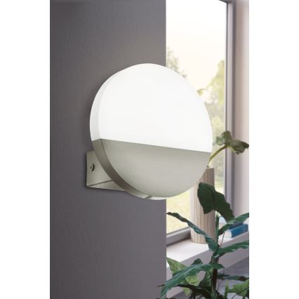 Okrúhla nástenná lampa svieti. Je pripevnená na tmavosivej stene. V pozadí je vidno okno a rastlinu v kvetináči.