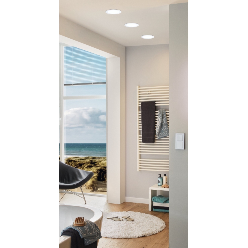 Tri vstavané svietidlá osvetľujú modernú kúpeľňu. Na stene je umiestnený elektrický sušiak na uteráky s viacerými uterákmi. V pozadí je vidieť výhľad na more cez veľké okno.