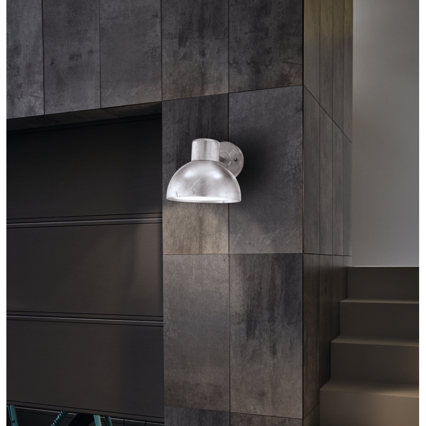 Strieborná nástenná lampa s polguľovitým tienidlom svieti na tmavosivej kachličkovej stene. Lampa je pripevnená k stene vedľa tmavej garážovej brány a schodiska.