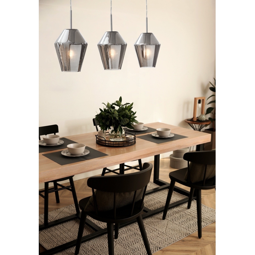 Tri visiace svietidlá s dymovým skleneným tienidlom v tvare mnohouholníka osvetľujú jedálenský stôl s drevenou doskou a čiernymi stoličkami. Stôl je prestretý.