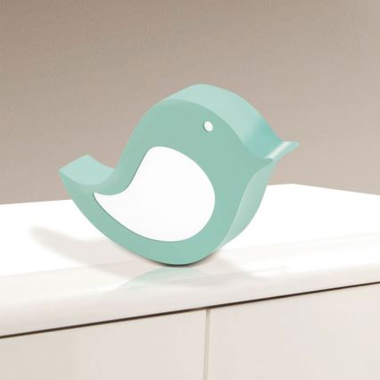 Drevená vtáčia lampa v svetlomodrej farbe stojí na bielom nábytku. Svietiaca časť lampy je biela.
