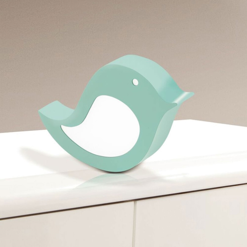 Drevená vtáčia lampa v svetlomodrej farbe stojí na bielom nábytku. Svietiaca časť lampy je biela.