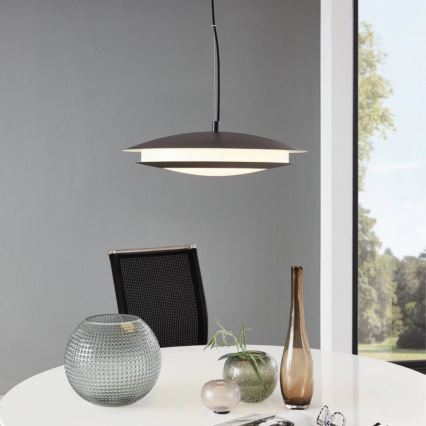 Závesné svietidlo svieti nad stolom. Svietidlo má tmavosivý kovový kryt a svetlý difúzor. Na stole sú vázy a stolová lampa.