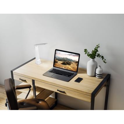 Stolová lampa svieti na pracovnom stole. Na stole je notebook, mobilný telefón a vázy s rastlinami. Pracovný stôl je drevený s čiernymi kovovými nohami.