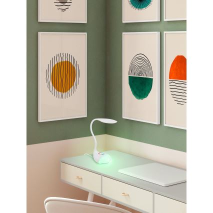 Biela stolová lampa s flexibilným ramenom svieti na pracovnom stole. Lampa vyžaruje zelenkavé svetlo. Na stene visia obrázky s geometrickými vzormi.