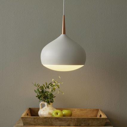 Svetlosivá závesná lampa s medeným detailom svieti. Pod ňou je na drevenom podnose váza s kvetmi a zelené jablká. V pozadí je sivá stena.