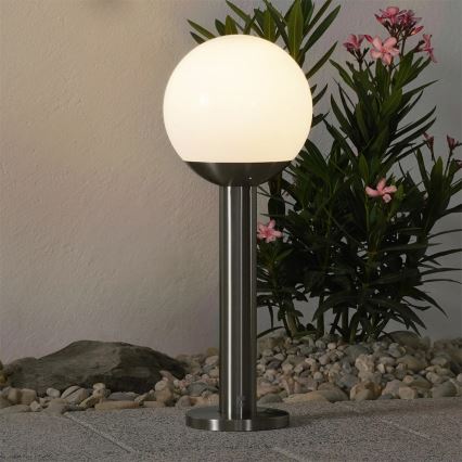 Svietidlo s guľovou tienidlom svieti. Stojí na kovovom stojane v exteriéri, medzi kameňmi a rastlinou s ružovými kvetmi.