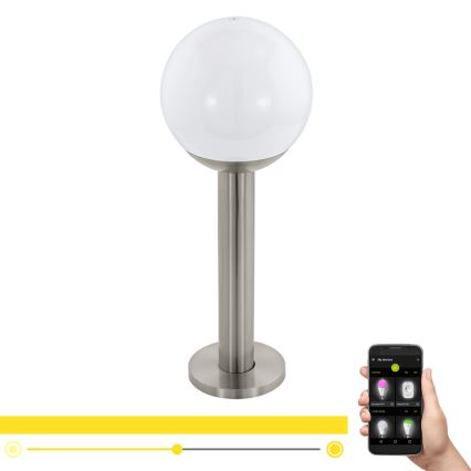 Svietidlo s bielou guľovou hlavou a kovovou nohou stojí na podstavci. Ovládanie prebieha cez mobilnú aplikáciu zobrazujúcu možnosti nastavenia farieb osvetlenia.