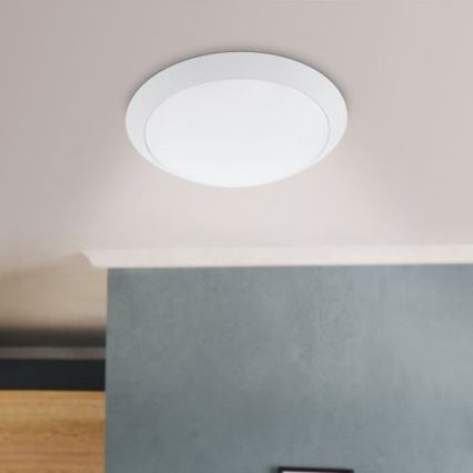 Svetlo je namontované na svetlom strope. Svietidlo je okrúhle, biele s tenkým bielym rámom. Pod ním je sivá stena a časť drevenej nábytku.