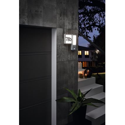 Svietidlo s číslom 78b je namontované na stene vedľa garážových dverí. Svietidlo svieti v noci. V pozadí sa nachádzajú schody a okolitá záhradná architektúra.