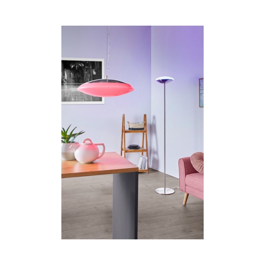 Zavesená lampa svieti ružovým svetlom v interiéri. Stojacia lampa svieti v blízkosti pohovky. Drevený stôl s čajovou kanvicou je osvetlený zavesenou lampou.