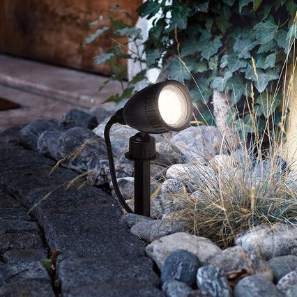 Záhradné svietidlo svieti. Je umiestnené v kamenistej záhradke, medzi okrasnými rastlinami.