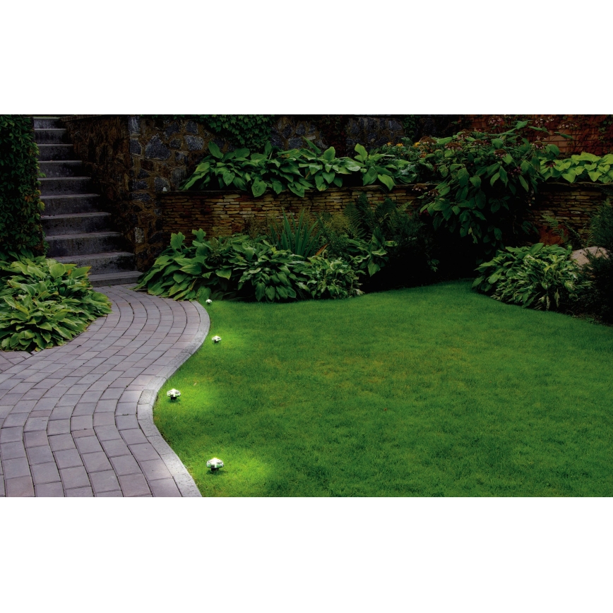 Záhradné svietidlá osvetľujú dlažbovú cestu popri trávniku. Svietidlá sú umiestnené pozdĺž okraja chodníka. Okolím je zeleň, kríky a kamenná stena.