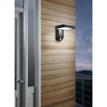 Čierne vonkajšie nástenné svietidlo s pohybovým senzorom je pripevnené na drevenej stene vedľa dverí. Svietidlo svieti. Na pozadí je vidieť nočnú oblohu a zábradlie.