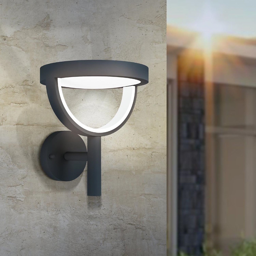 Vonkajšie nástenné svietidlo svieti na betónovej stene. Svietidlo je tmavosivé s bielym vnútorným kruhom. Je namontované na stene v exteriéri budovy.