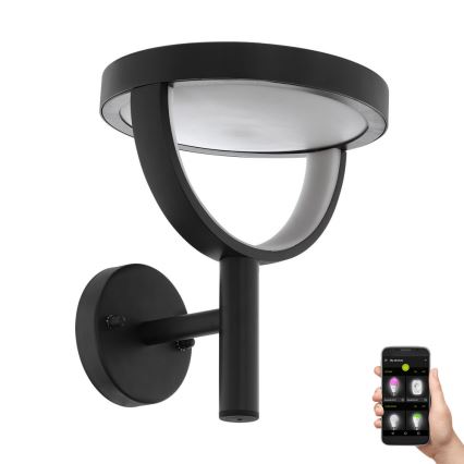 Čierna nástenná lampa s okrúhlou tienidlom svieti. Lampa je ovládaná mobilnou aplikáciou zobrazenou na smartfóne, ktorá umožňuje meniť farebné nastavenie svetla.