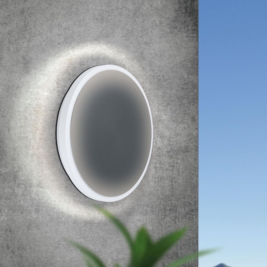 Okrúhle nástenné svietidlo svieti na betónovú stenu. Svietidlo má sivý stred a biely rám. Na pozadí je vidieť modrú oblohu a časť zelenej rastliny.