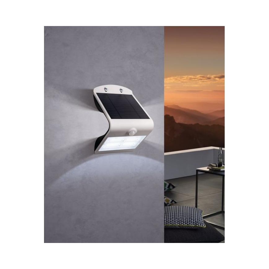 Solárny senzorový nástenný svietidlo je pripevnené na stene. Svietidlo svieti. Na pozadí je vidieť západ slnka a terasa.