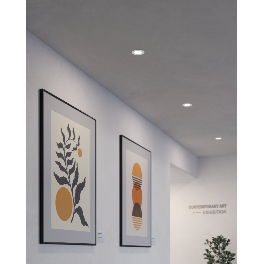 Dve obrazy s abstraktnou grafikou visia na stene. Svietidlá sú zabudované do stropu. Na stene je nápis: „CONTEMPORARY ART EXHIBITION“.