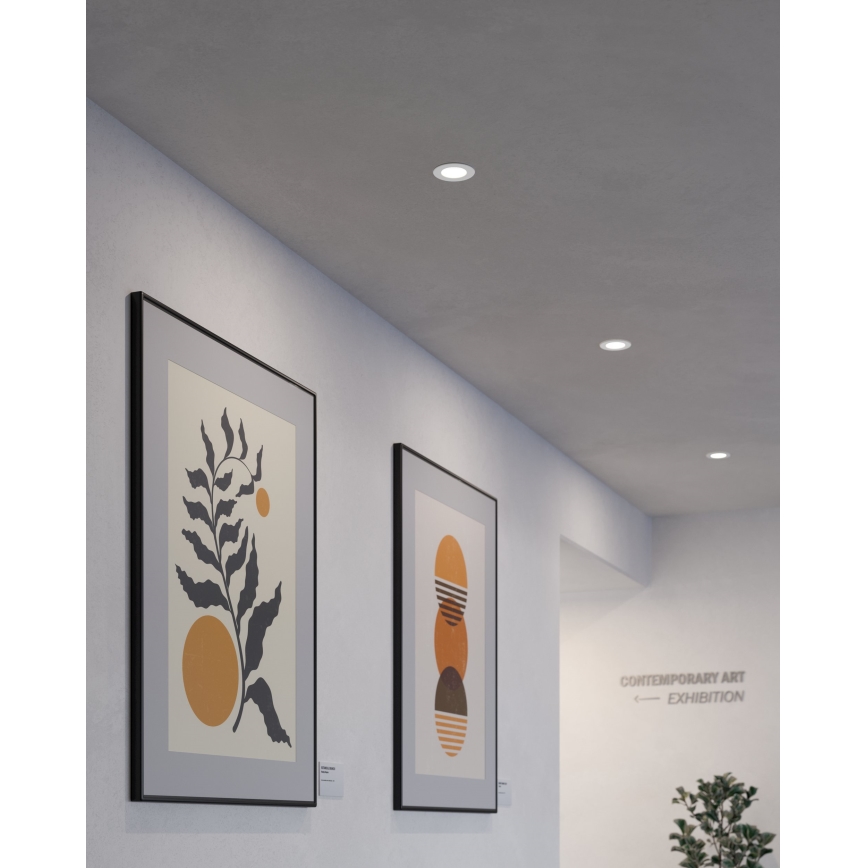 Dva obrazy s abstraktnou geometrickou a botanickou tematikou visia na stene v chodbe. Na stene je označenie „CONTEMPORARY ART EXHIBITION“. Svietidlá sú zabudované do stropu.