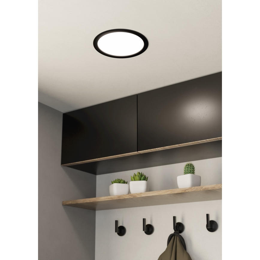 Okrúhle stropné svietidlo svieti v interiéri. Nachádza sa nad drevenou poličkou s kvetináčmi a háčikmi na stene. Polička je umiestnená pod tmavými skrinkami.