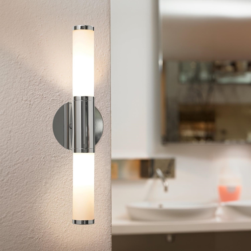 Nástenná lampa svieti na svetlú stenu, s chrómovými detailmi. V odraze je viditeľný umývadlový priestor s kohútikom a pohárom s ružovou tekutinou.